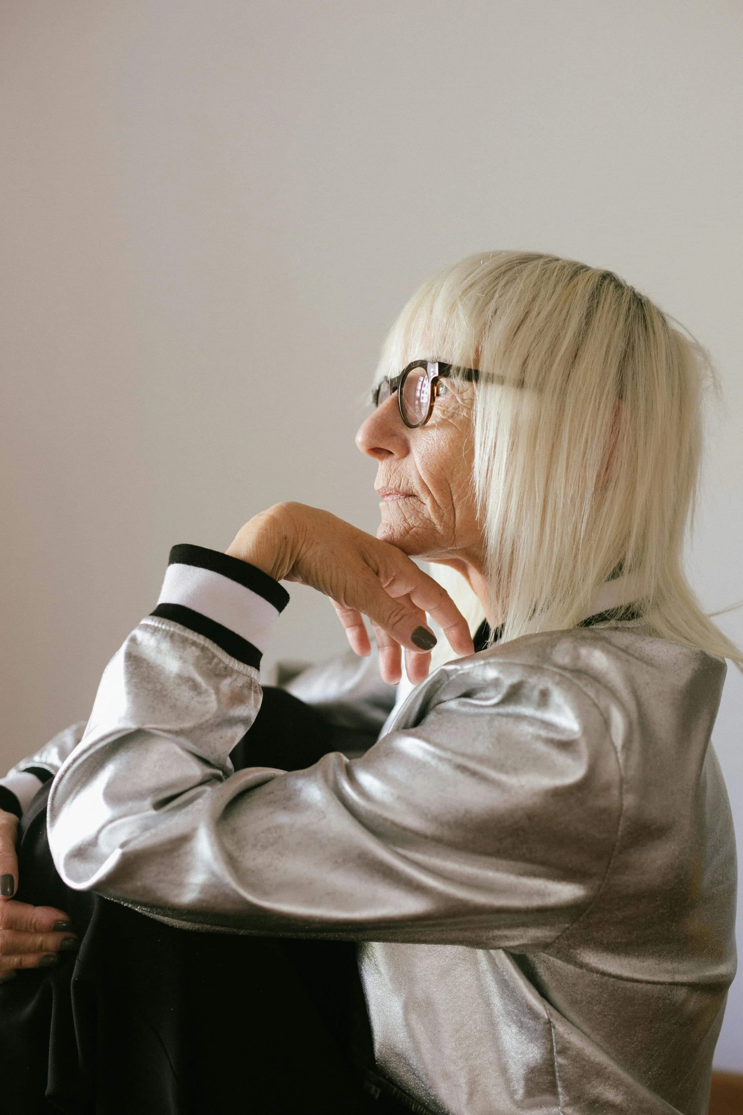 A stylish elderly woman wearing glasses and a silver jacket, looking off to the side.