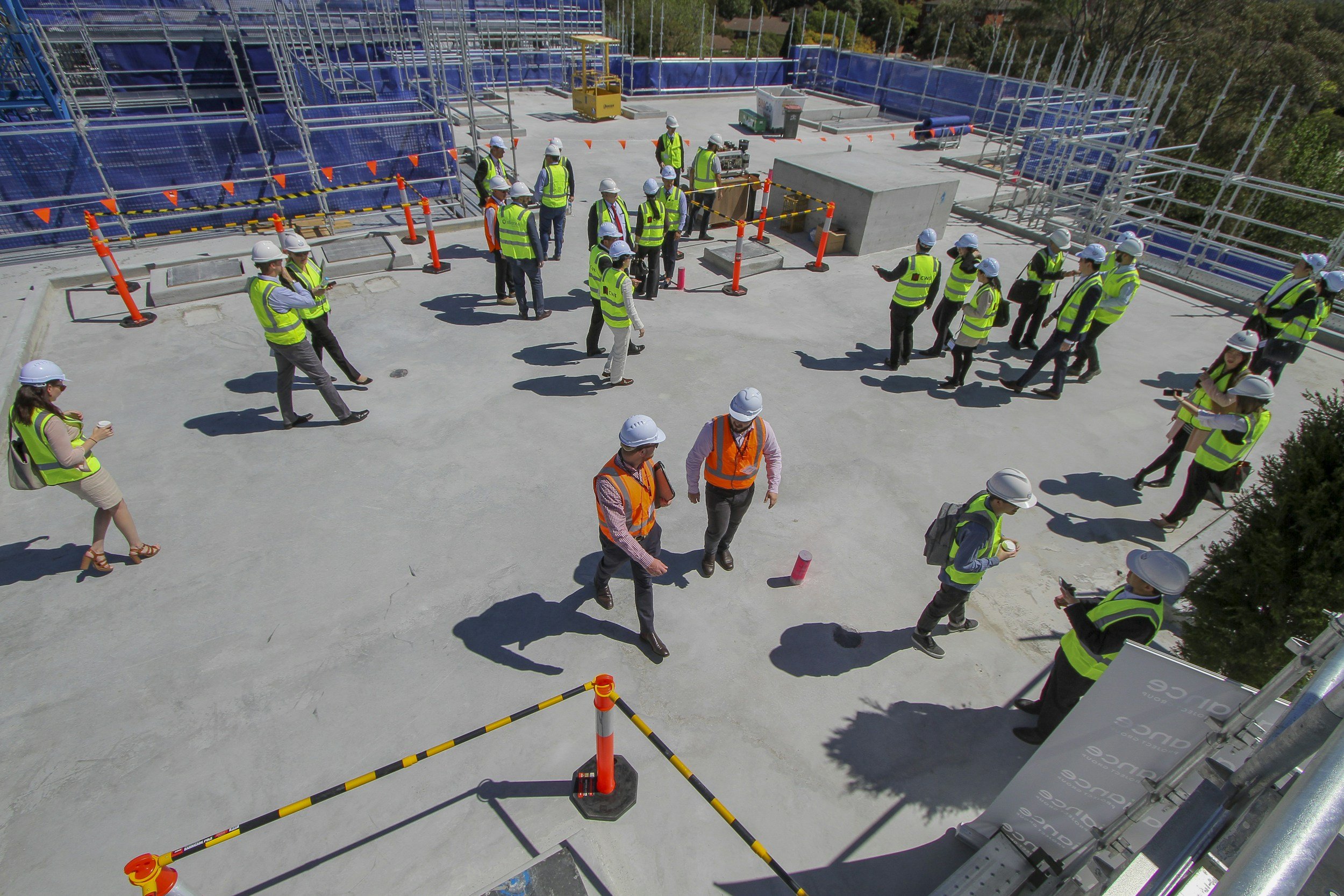 A group of construction workers and officials wearing safety vests and helmets on a construction site, with some standing near orange cones and barriers, and scaffolding in the background.