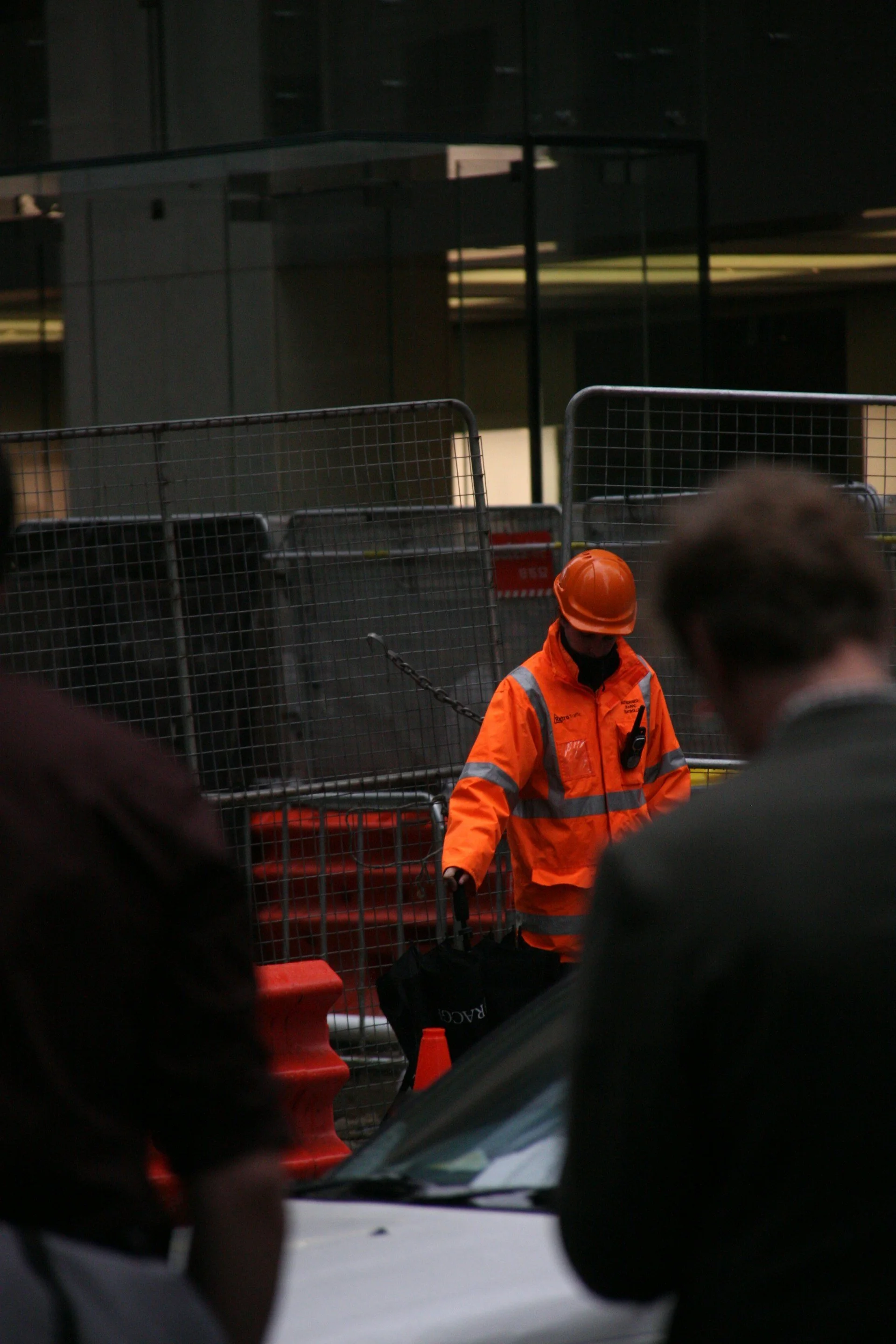 A construction worker dressed in an orange safety jacket and hard hat, standing behind a chain link fence, holding a black bag, with blurred people and a black car in the foreground.