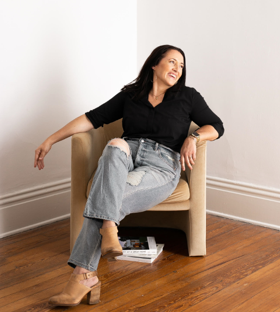 Woman sitting on an armchair smiling and looking to her left, wearing a black shirt, ripped jeans, and tan high-heeled shoes, with a magazine under her feet, in a room with white walls and hardwood floor.