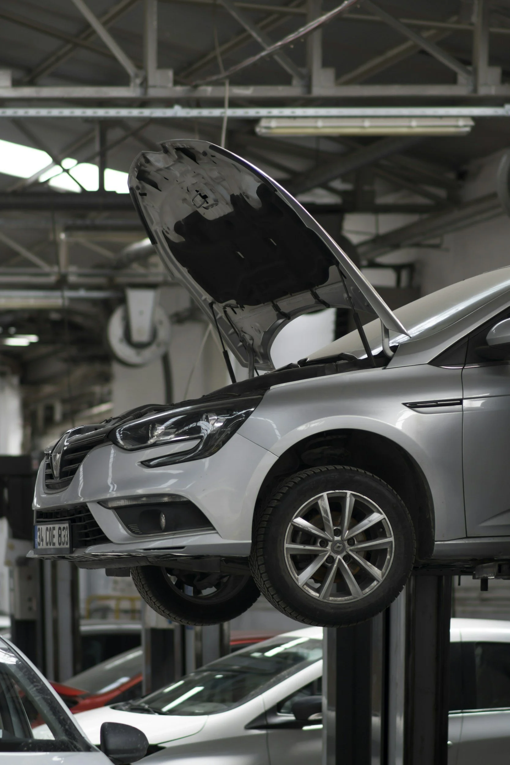 A silver car is lifted on a mechanic’s lift inside an auto repair shop, with its hood open for maintenance or inspection.
