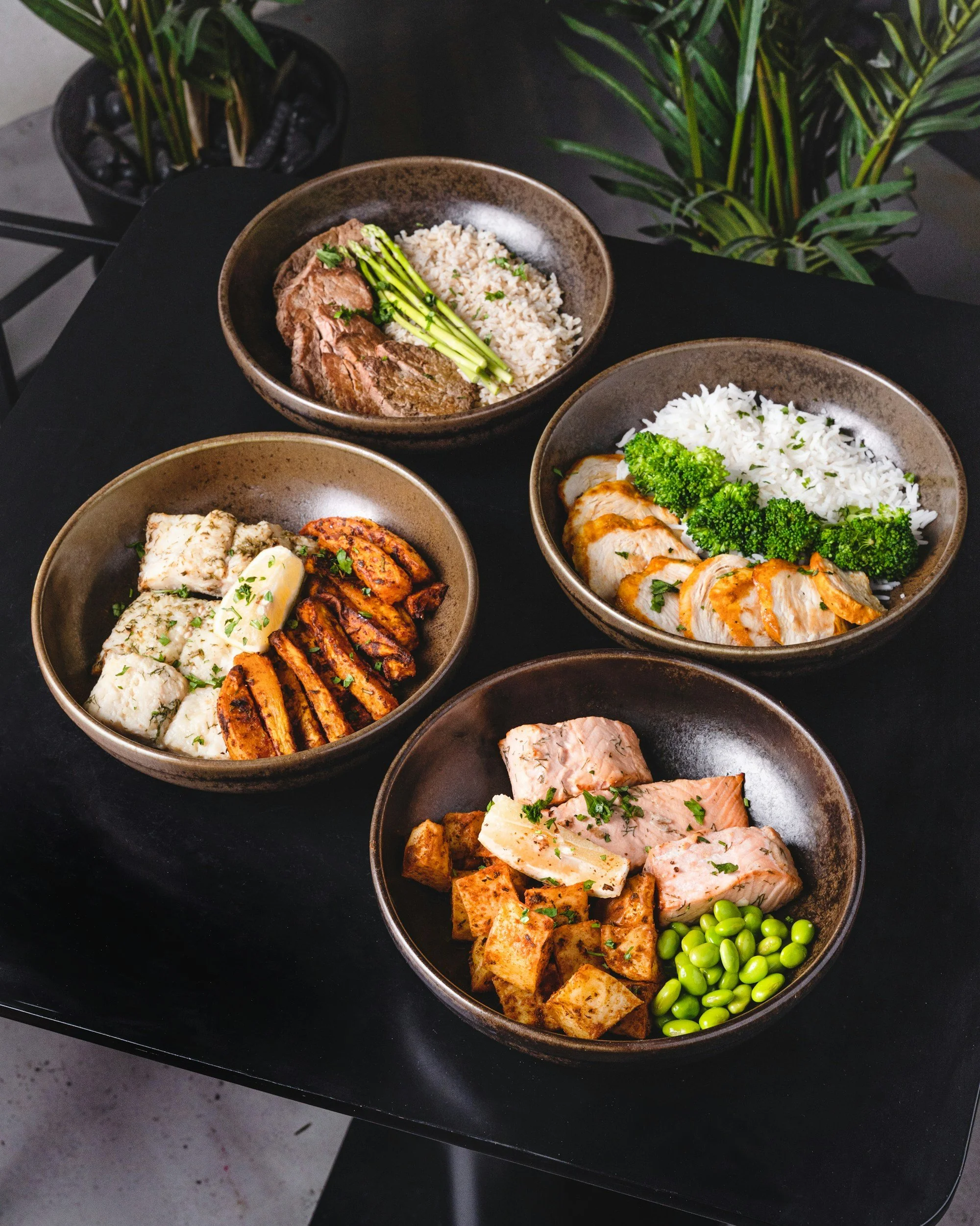Four bowls of food on a black table, each containing different meals with rice, grilled chicken, beef, fish, grilled vegetables, and tofu, with green garnish.