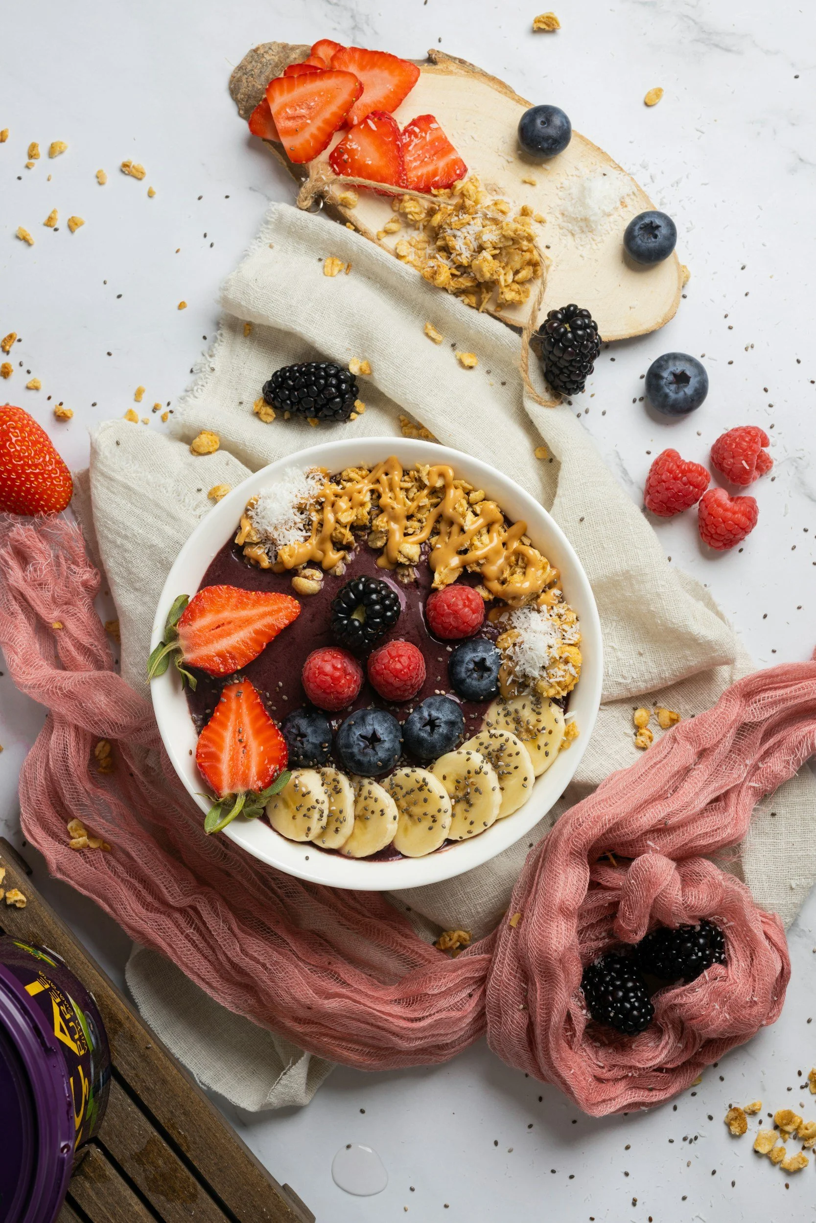 Bowl of smoothie topped with strawberries, blackberries, raspberries, banana slices, shredded coconut, and granola on a white surface with scattered berries and crumbs, surrounded by cloth napkins and a wooden board.