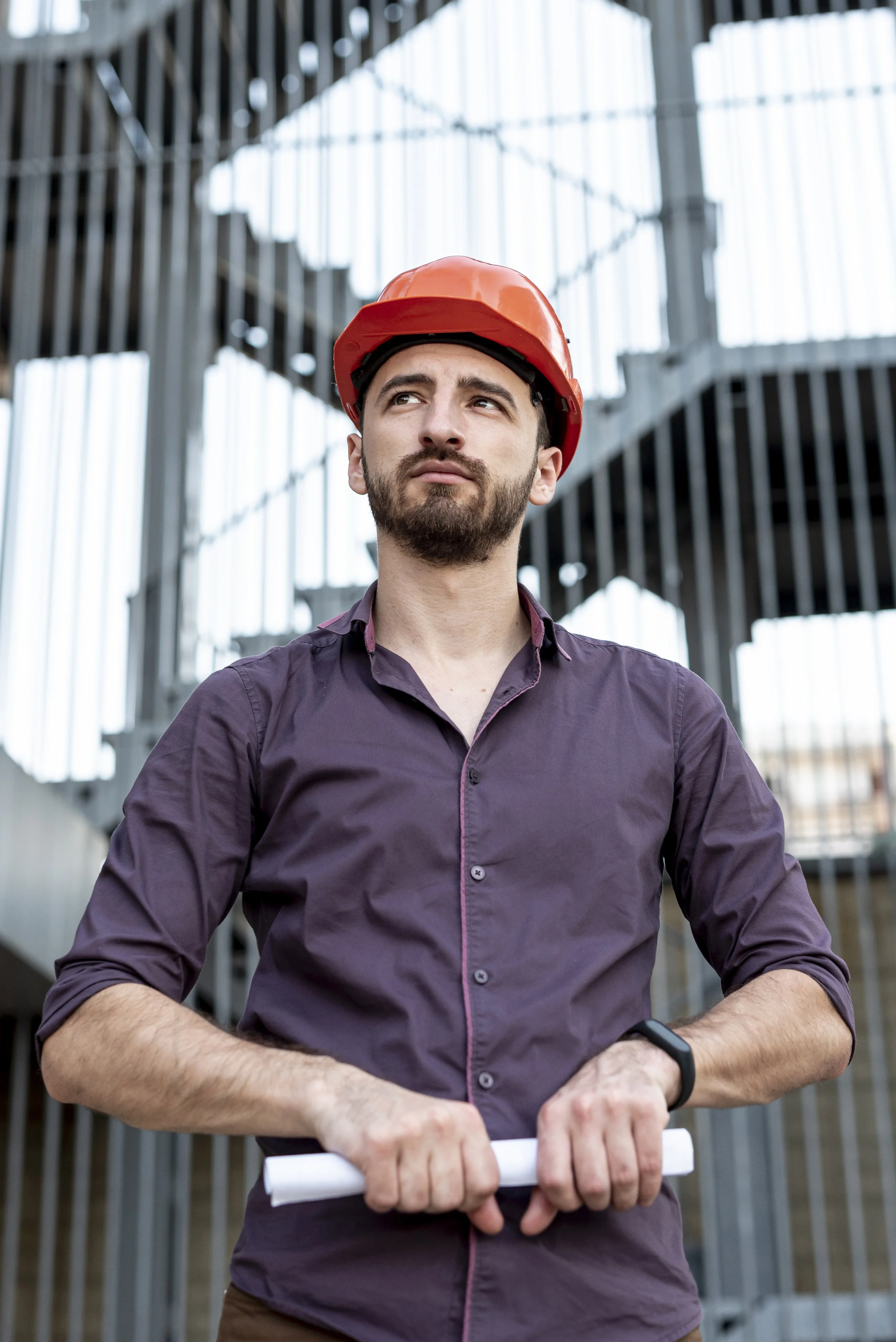 A man with a beard and mustache, wearing a red safety helmet and a purple button-up shirt, holds rolled-up blueprint plans and looks thoughtfully into the distance at a construction site with metal framework background.