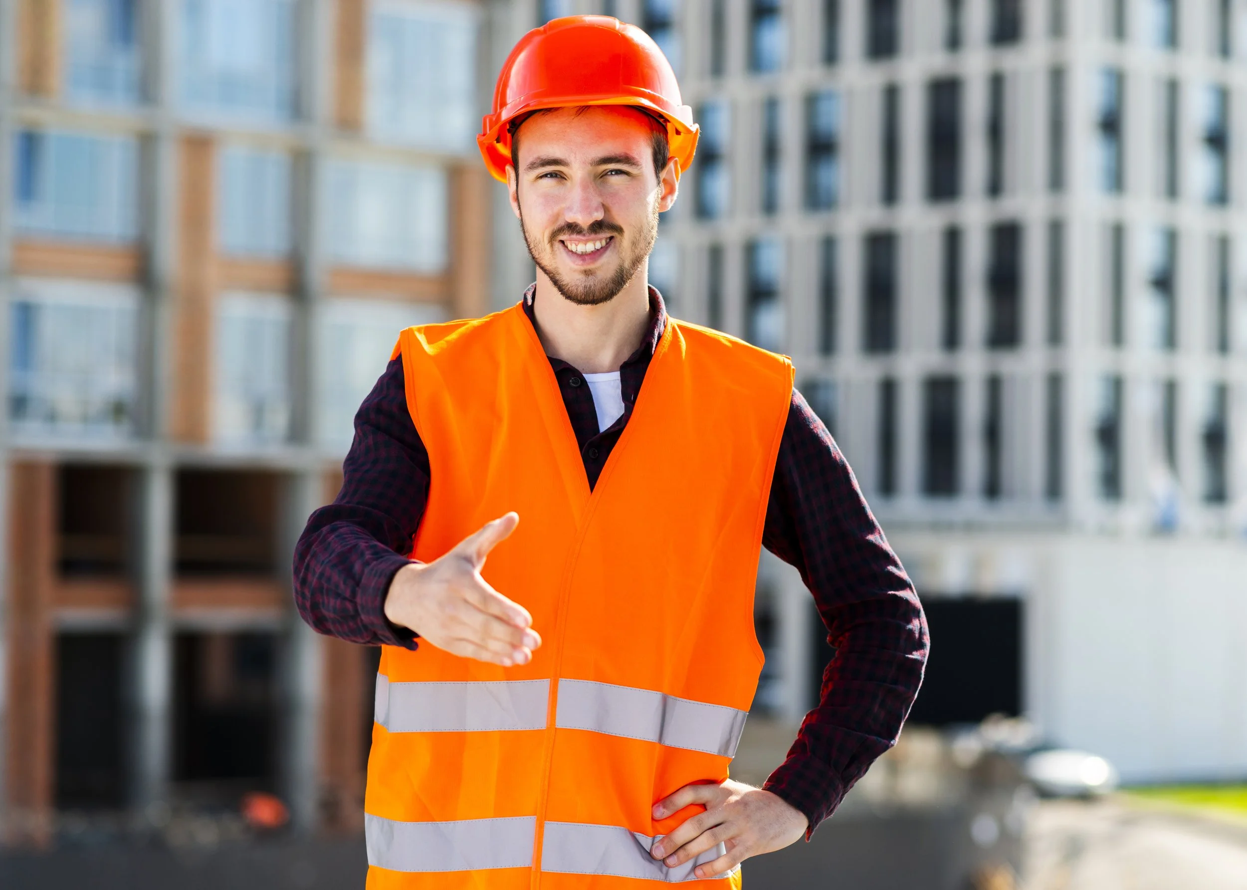 A construction worker smiling and shaking hands on a construction site, wearing an orange safety vest and a red safety helmet.
