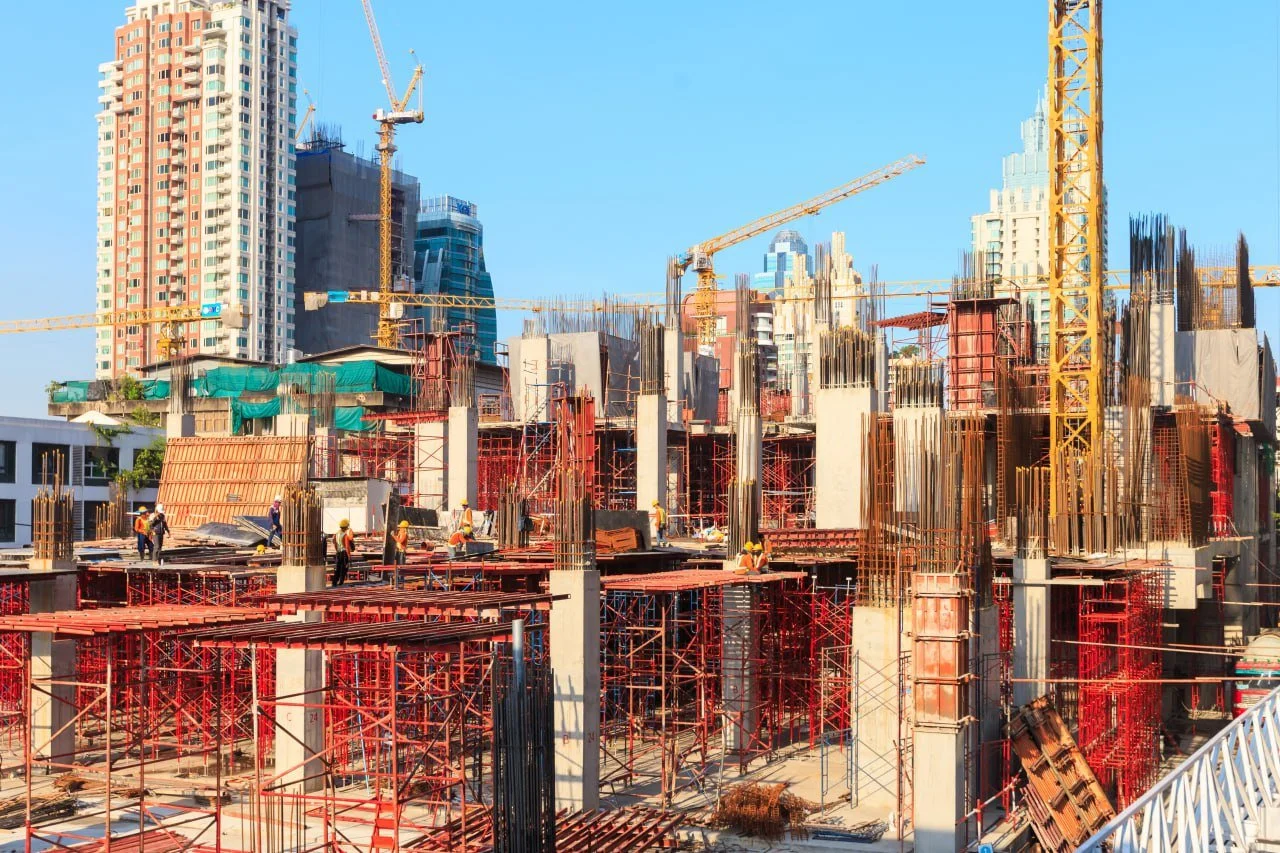 Construction site with workers, cranes, and steel framework, with high-rise buildings in the background.
