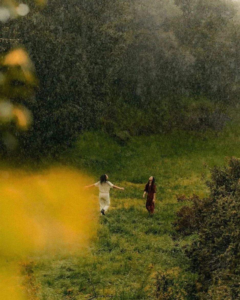 Two women are walking in a grassy field at night, holding hands, with trees and dark sky in the background.