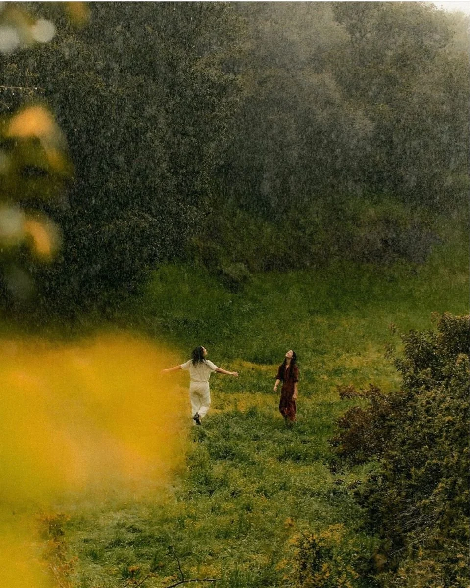 Two women walking through a grassy field surrounded by trees on a cloudy day.