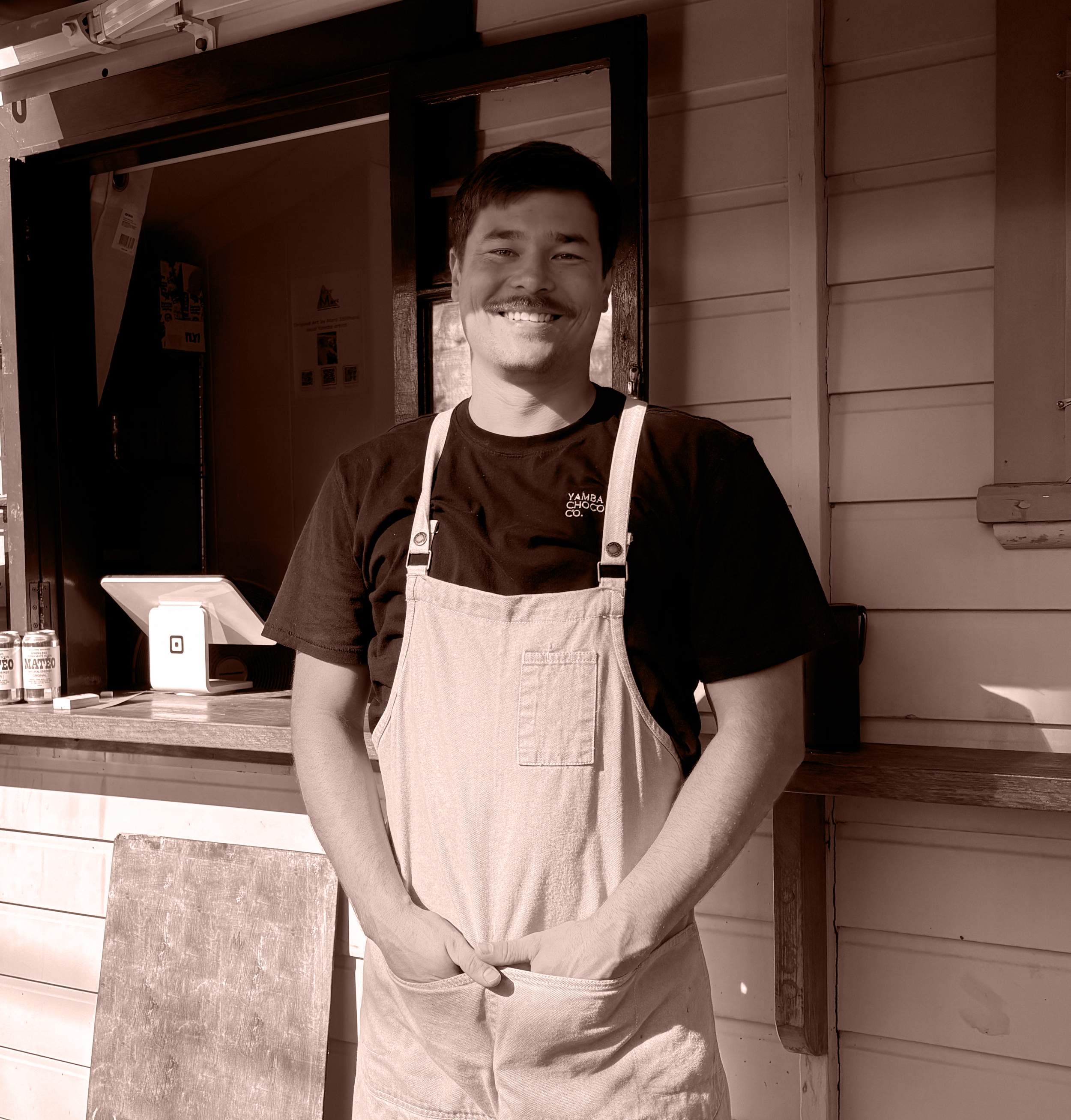 Kieran, Yamba Chocolate Co's founder and chocolate maker wearing an apron and smiling while standing behind a counter in a rustic, wooden setting.