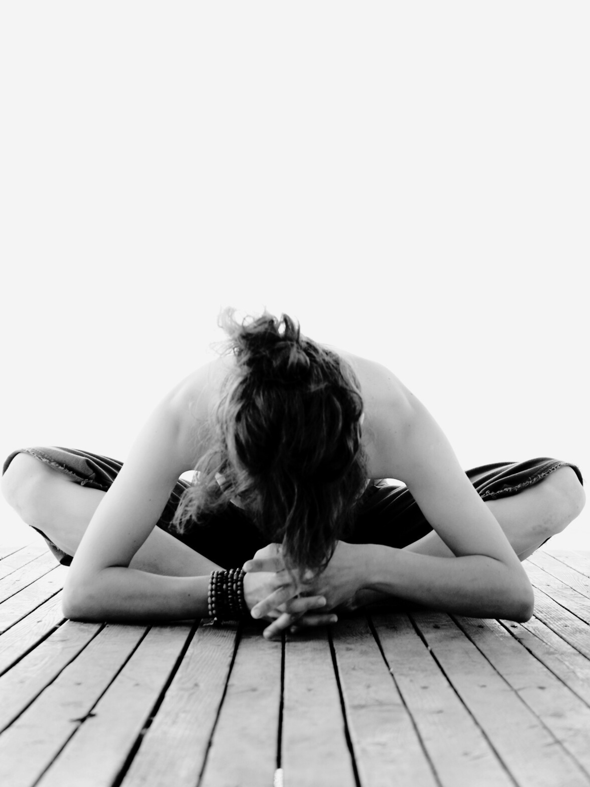 Person practicing yoga on a wooden floor, sitting cross-legged with hands clasped in front of face, head bent down, in a pose of meditation or stretching, wearing bracelets and loose clothing.