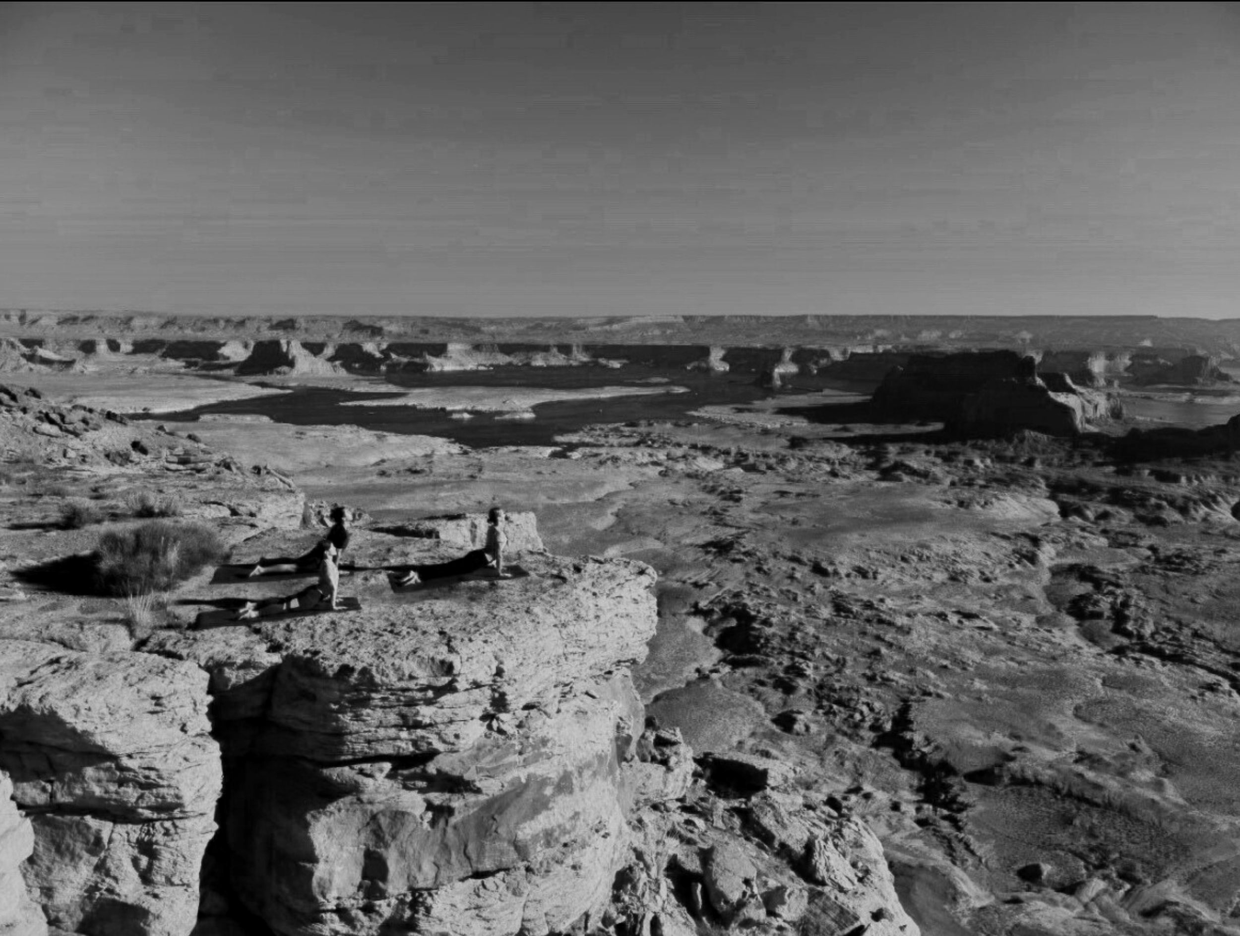 Black and white photo of a desert landscape with rocky terrain, mesas, and canyons in the distance, with two people practicing yoga on a large flat rock in the foreground.