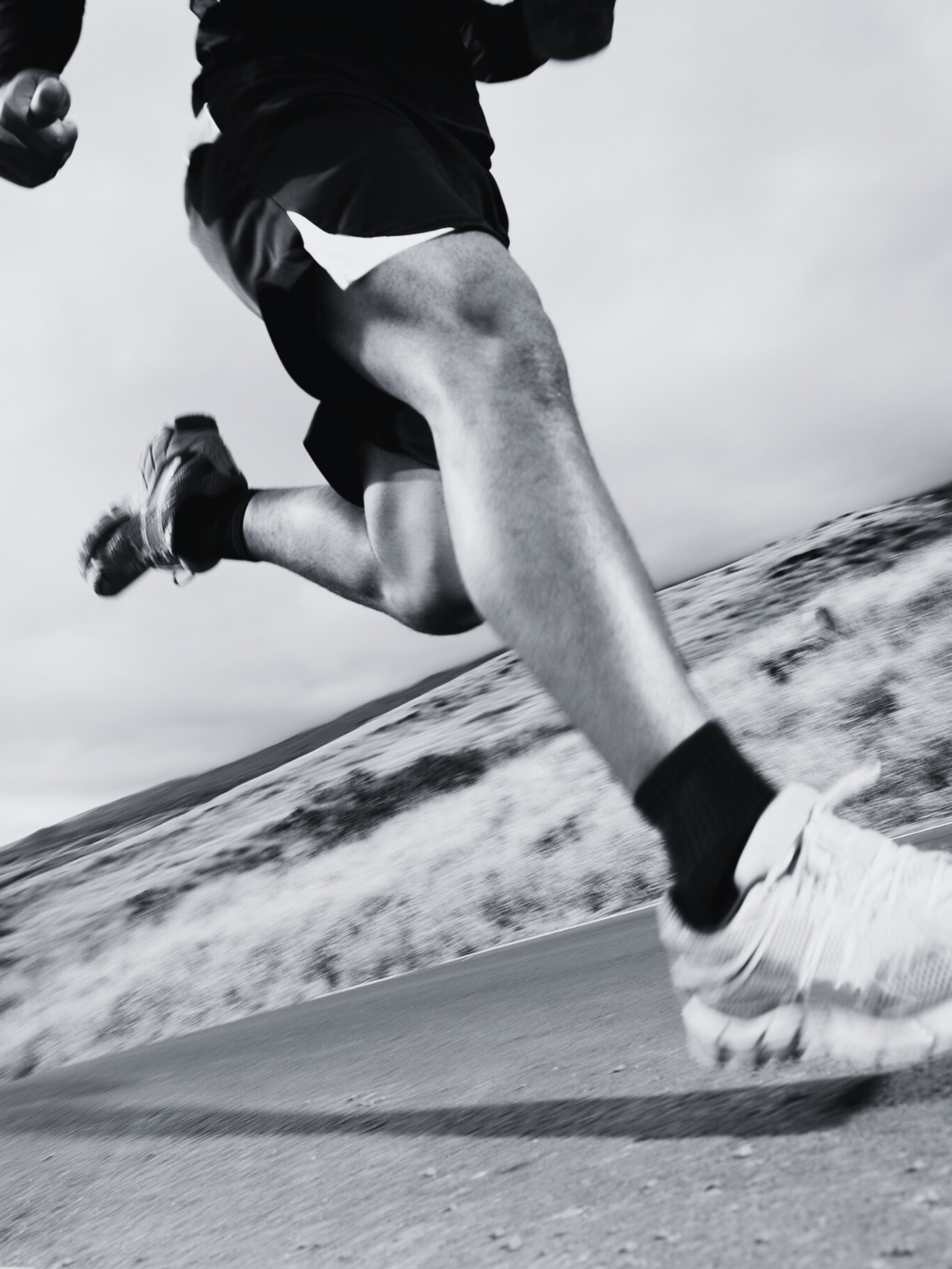Close-up of a person running outdoors on a paved road, wearing athletic shorts, running shoes, and black socks, with a blank sky and hilly landscape in the background.