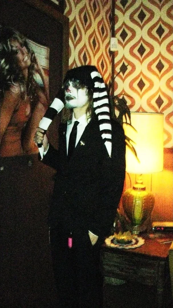 Person dressed as a mime with black and white striped hair, wearing a black suit, standing beside a lamp and a patterned wall.