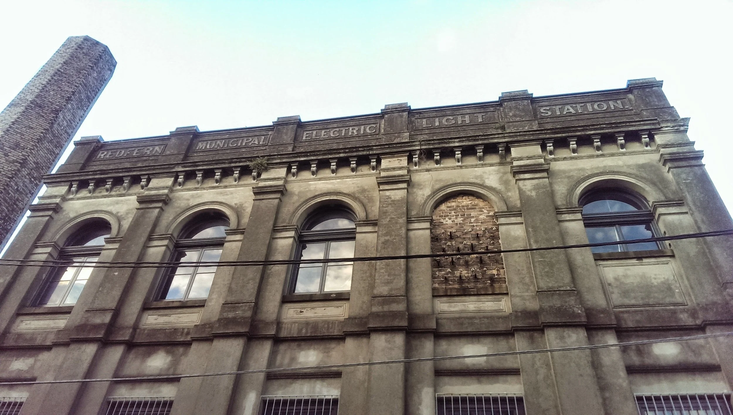 Old brick building with arched windows and a sign on top that reads 'Redfern Municipal Electric Light Station', with some wires crossing in front.
