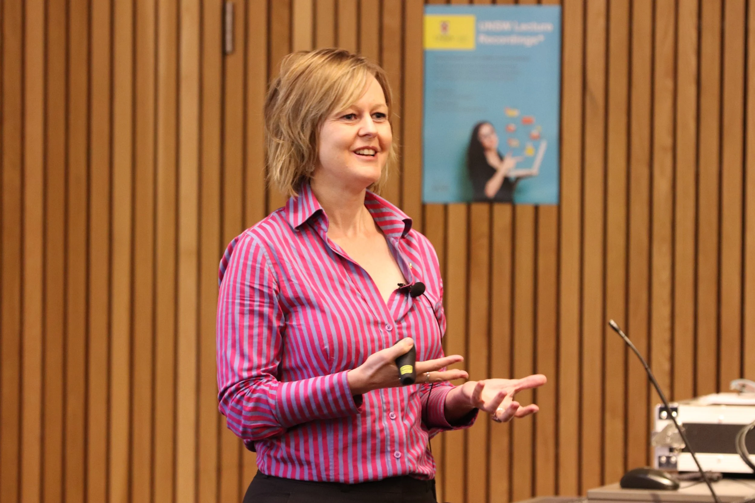 Woman giving a presentation in a conference room with wooden walls, holding a clicker, standing next to a desk with electronic equipment.
