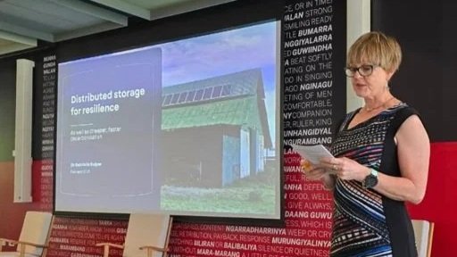 Gabrielle Kuiper speaking at the Neighbourhood Battery Summit at ANU in Canberra with a large display screen showing a presentation slide titled 'Distributed storage for resilience'.