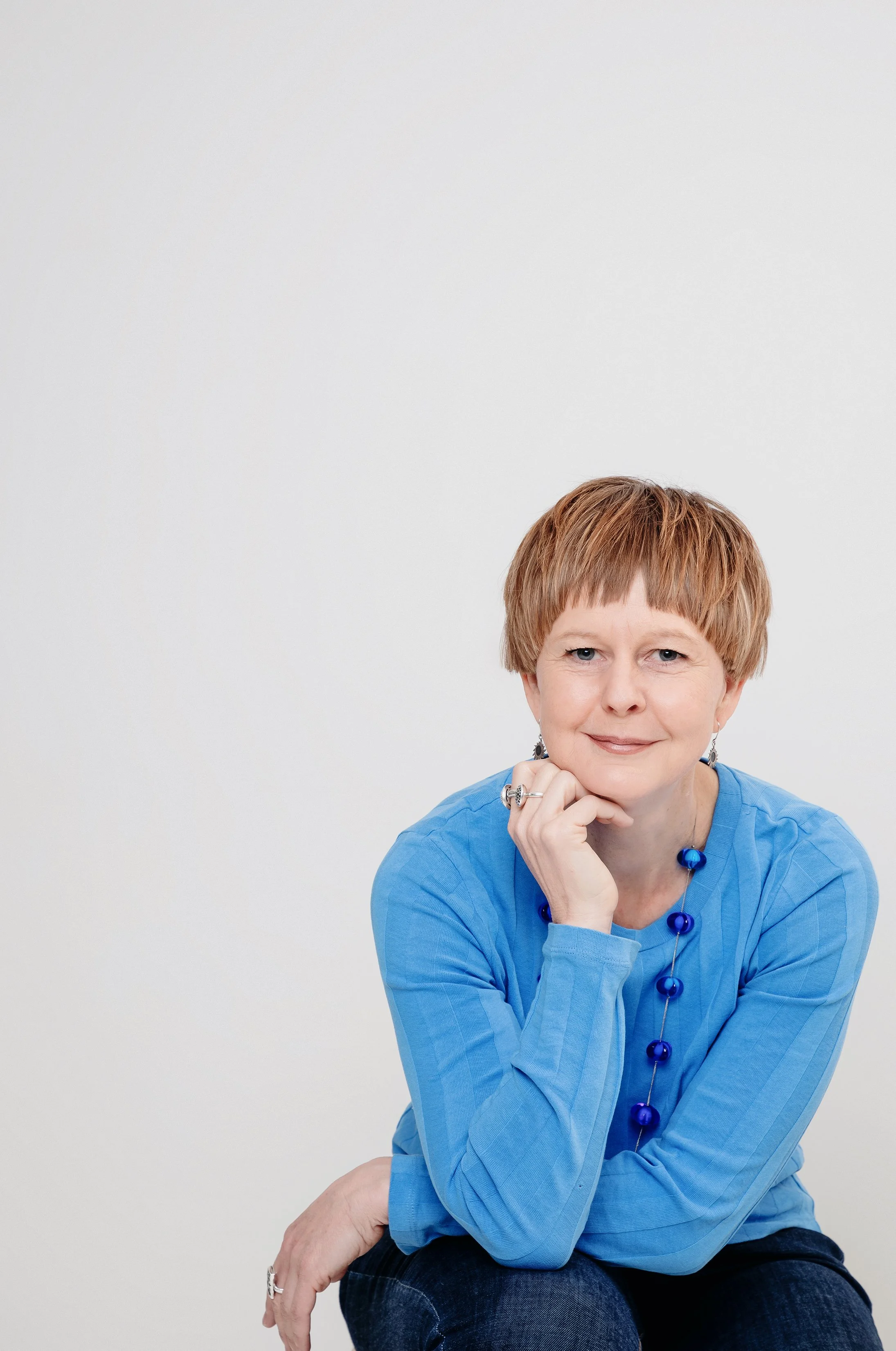 Professional portrait Gabrielle Kuiper with short brown hair and blue eyes smiling, resting her chin on her hand, wearing a blue top and matching blue jewellery, sitting against a plain white background.