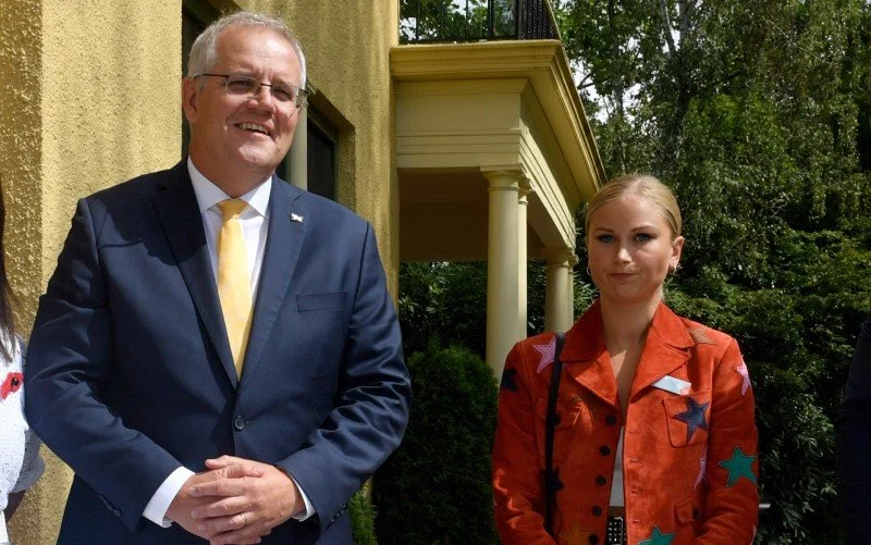 Former Prime Minister Scott Morrison a navy suit and yellow tie standing outdoors next to a Grace Tame in an orange jacket with stars, with a yellow building and lush green trees in the background.