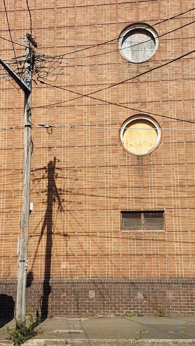Brick building wall with two circular vents, an electrical pole with wires, and a shadow of the pole on the wall.
