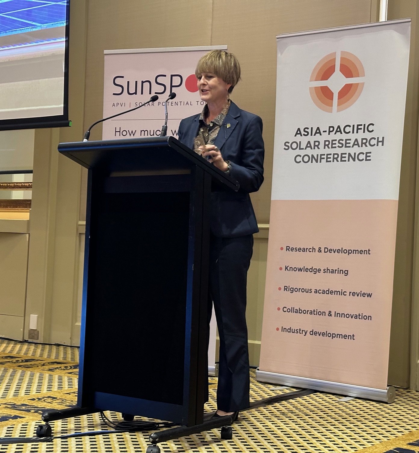 Dr Gabrielle Kuiper giving a keynote presentation at the Asia-Pacific Solar Research Conference, standing behind a black podium with a microphone. She is wearing a navy blazer and has short, light brown hair.