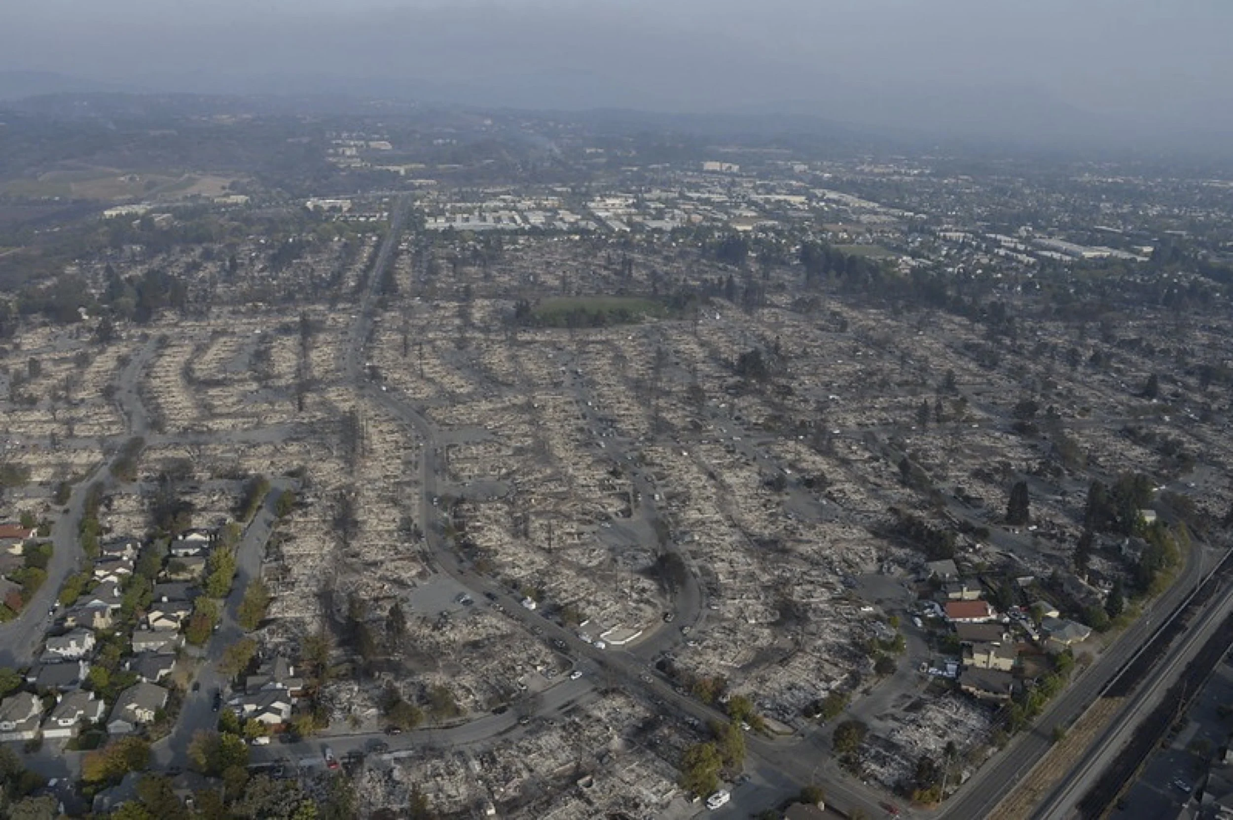 Aerial view of a neighborhood with many destroyed houses and burnt debris, likely from a recent fire, with some undamaged houses visible at the bottom left corner.