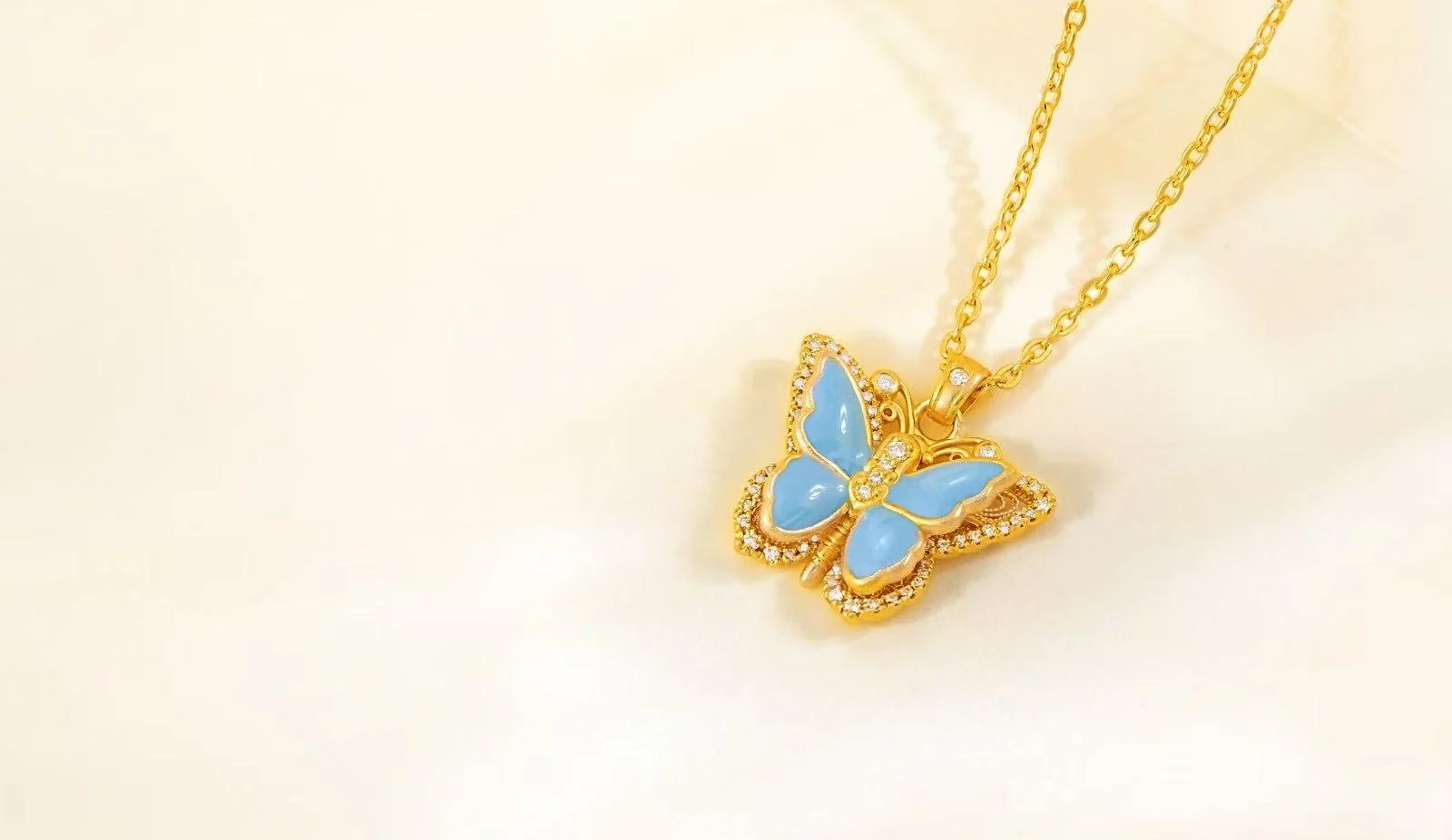 Gold butterfly necklace with light blue wings and small sparkling stones, hanging from a gold chain, on a light background.