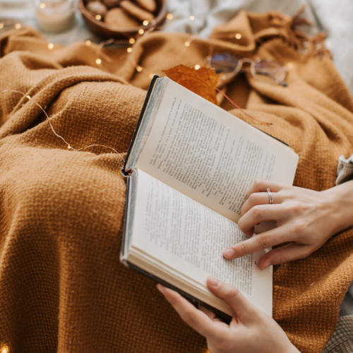A person reading a book while wrapped in a brown blanket, with cozy slippers and warm lighting in the background.