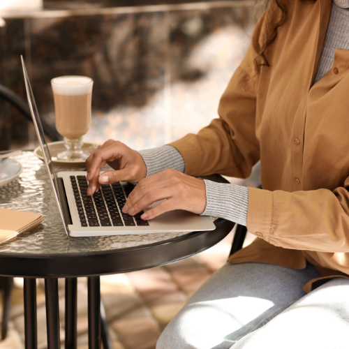 Person working on a laptop at an outdoor table, with a latte and a notebook beside them.