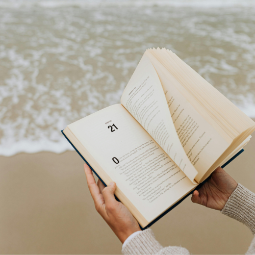 Person holding an open book near the beach with ocean waves in the background.