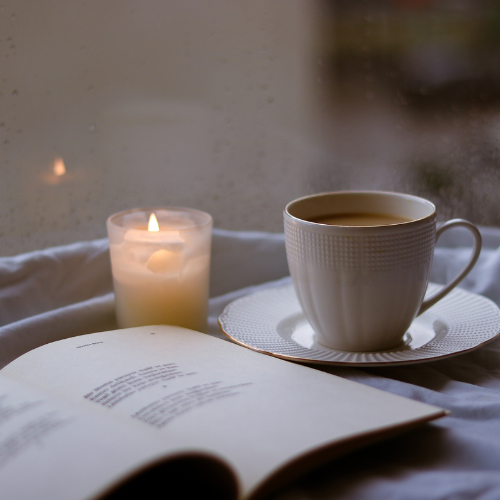 A white cup of coffee on a saucer, a lit candle, and an open book on a white surface near a window.