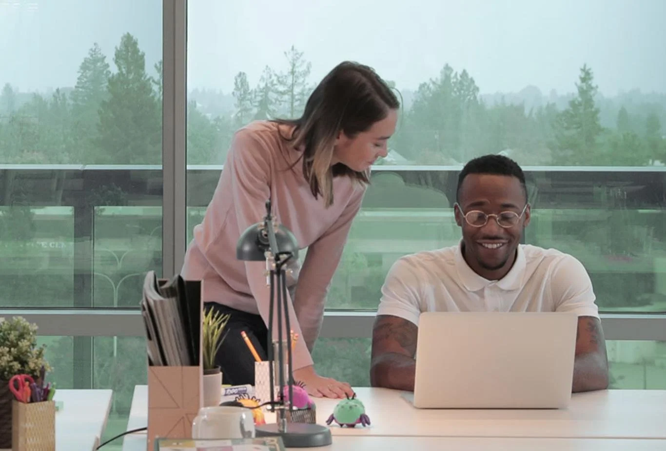 A woman and a man are working together at a desk in an office with large windows and a view of trees outside.