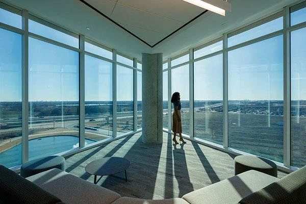 A woman standing in a high-rise room with floor-to-ceiling windows, overlooking a cityscape and a pool outside, in a modern interior with minimalist furniture.