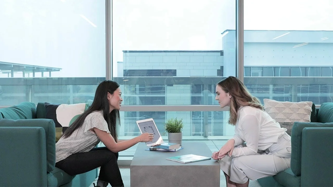 Two women sitting on teal armchairs across from each other in a modern office, engaged in a conversation. One woman is holding a tablet, and there are books and papers on the table between them, with large windows in the background.
