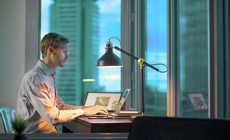 A man working on a laptop at a desk near a large window in an office, with a desk lamp, notebooks, and a framed photo.