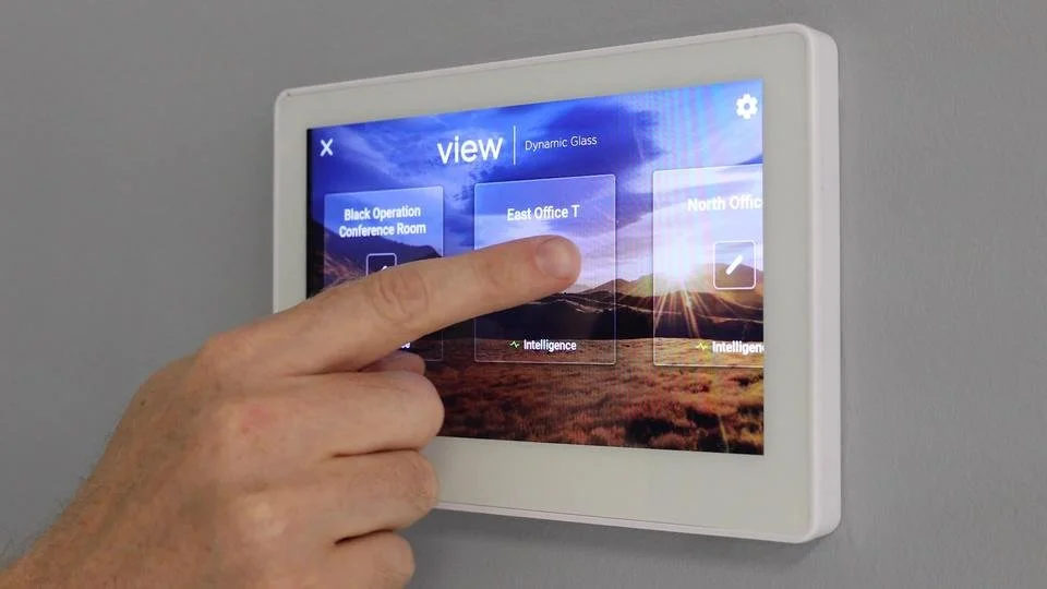 A person is touching a touchscreen control panel mounted on a wall, displaying options for managing different office conference rooms and areas.