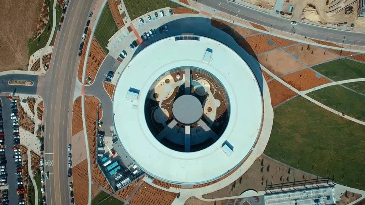 Aerial view of a large, circular building with a glass roof, surrounded by parking lots and green areas.