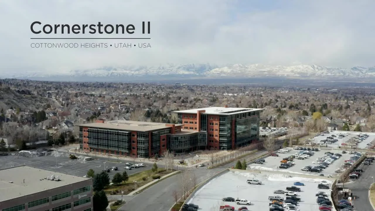 Aerial view of a modern office building with parking lots, in Cottonwood Heights, Utah, USA, with snow-capped mountains in the background.