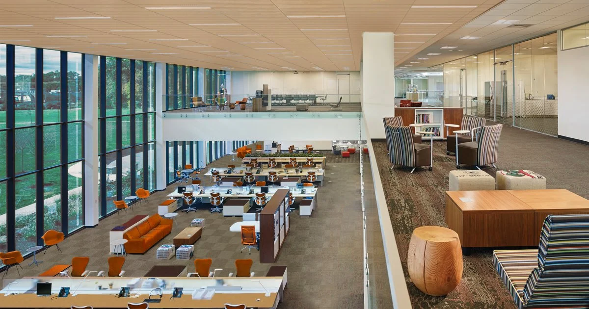 Interior of a modern office with large glass windows, open workspaces, and a separate lounge area with chairs and bookshelves.