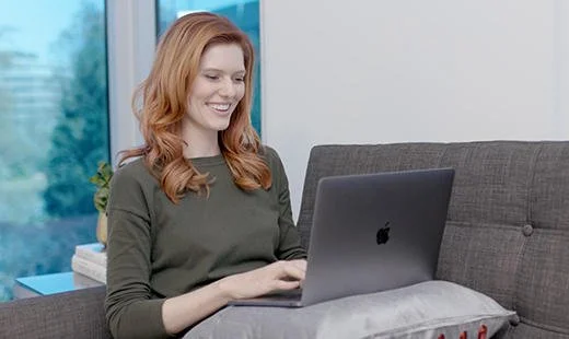 A woman with red hair sitting on a gray couch using a laptop, smiling, in a bright room with a large window.
