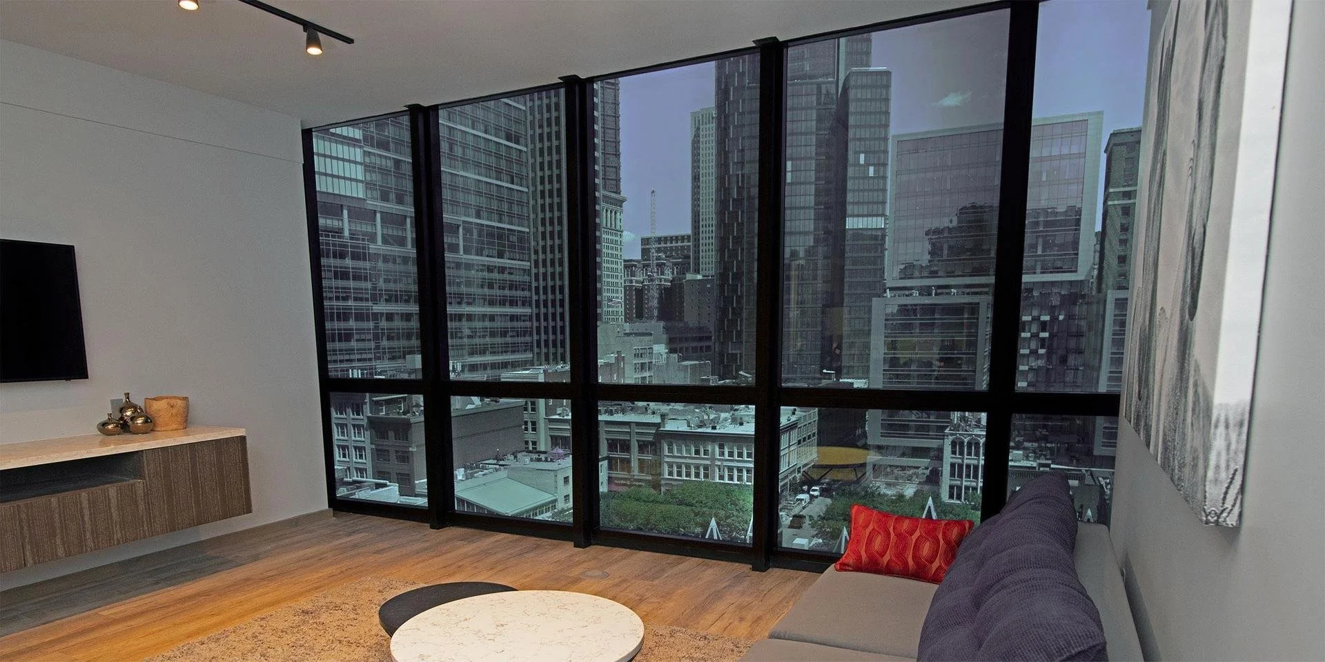 Modern living room with large floor-to-ceiling windows showcasing a city skyline of tall buildings, gray skies, and a few clouds. The room has a gray sofa with a red cushion, a round white marble coffee table, a wooden sideboard with decorative vases, and an abstract painting on the wall. The floor is wooden, and there is a beige rug.