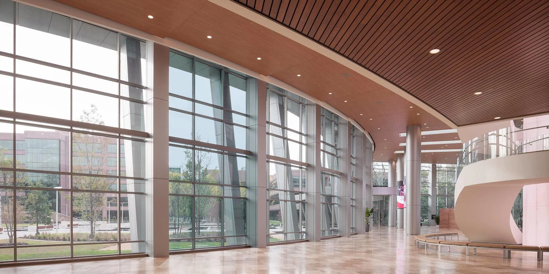 Modern building interior with large floor-to-ceiling glass windows, wooden ceiling, shiny metallic columns, and a curved white reception desk.