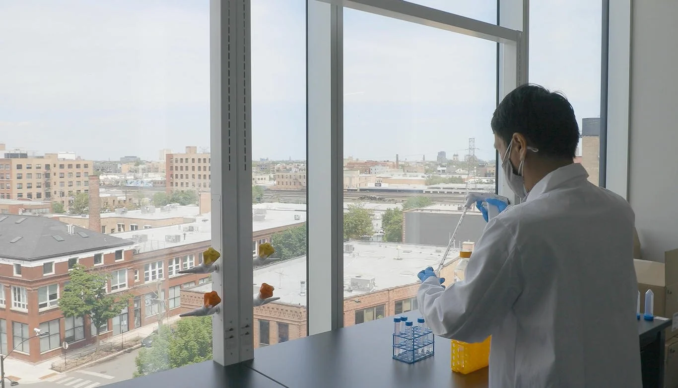 A scientist wearing a lab coat, face mask, and gloves conducts experiments using a pipette in a laboratory with large windows, overlooking a cityscape of buildings and streets.