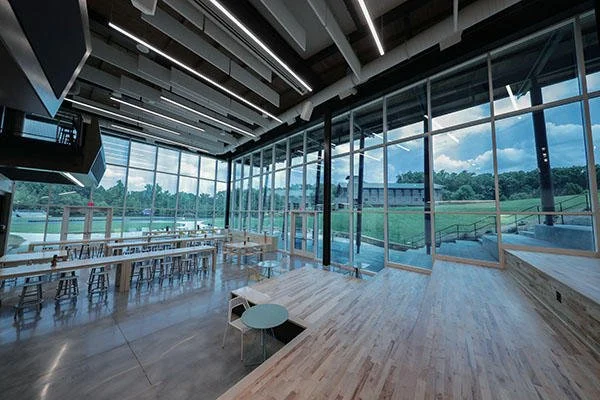Spacious, modern indoor space with large glass windows overlooking green outdoor scenery, featuring wooden flooring, tables, chairs, and an elevated platform or stage area.
