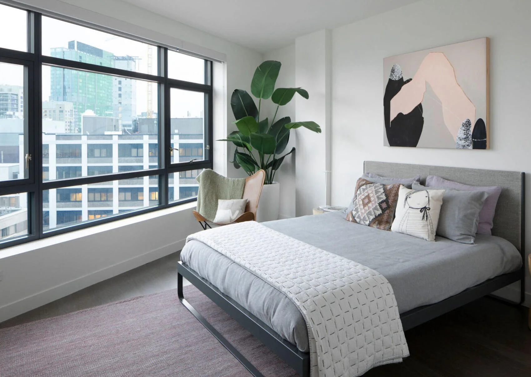 A modern bedroom with a large window overlooking a cityscape. The room features a bed with a gray upholstered headboard, decorated with patterned and plain pillows, and a white textured blanket at the foot. There is a tall green plant near the window and a wooden chair with a light green throw blanket and a pillow. Artistic wall decor hangs above the bed.