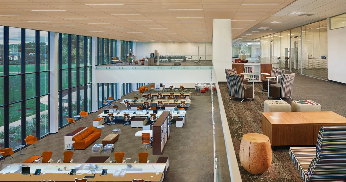 Modern office space with large windows, open work areas, and a separate lounge area with chairs and tables.