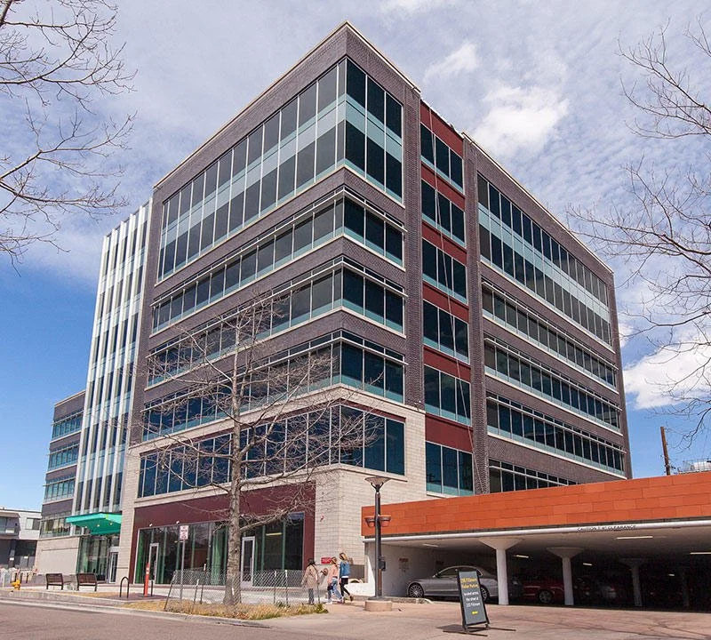 Modern multi-story office building with glass windows and a parking garage on the ground floor, trees and people in front, under a partly cloudy sky.