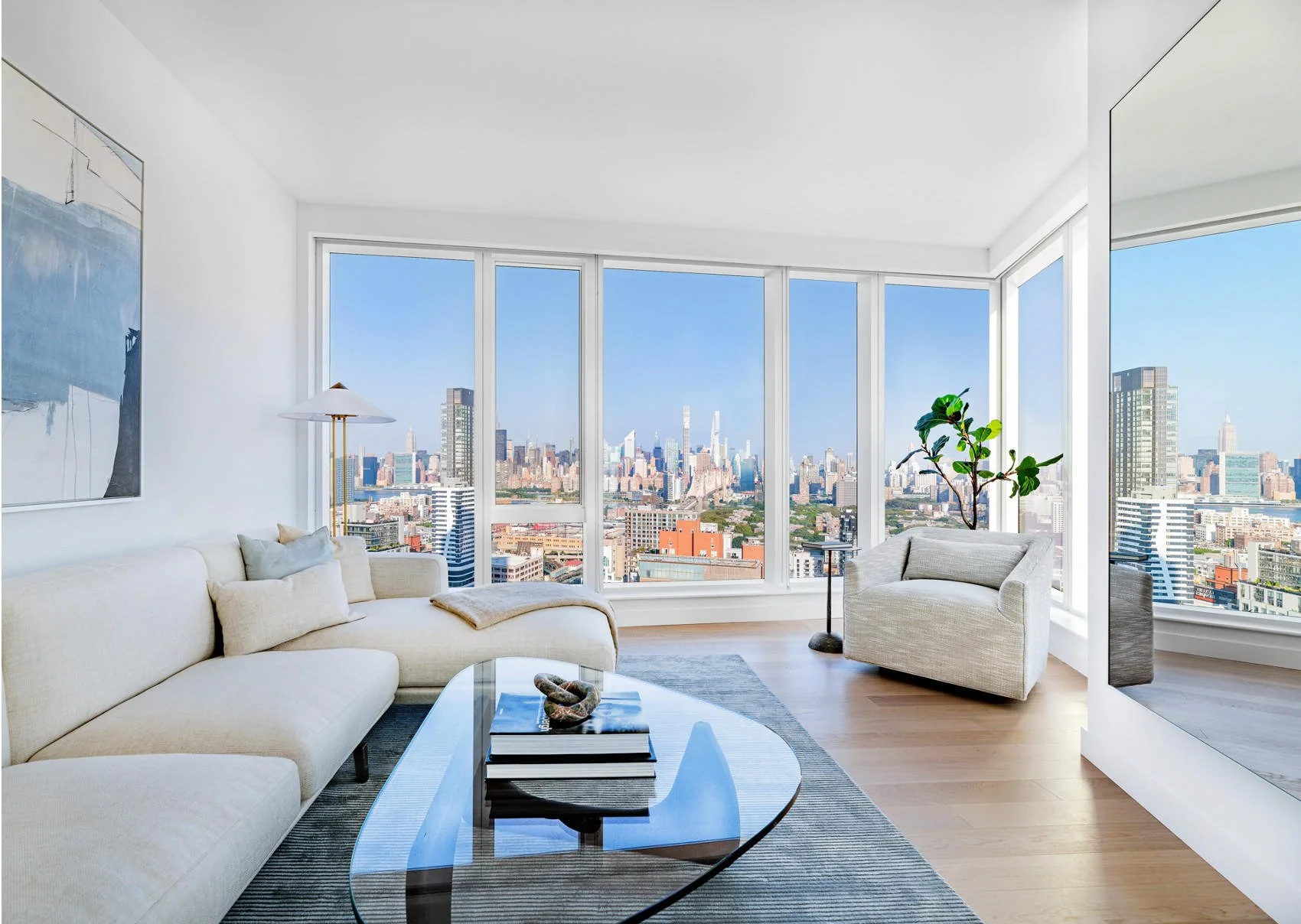Modern living room with panoramic city view through floor-to-ceiling windows, white sofa, armchair, glass coffee table, plants, and contemporary artwork.