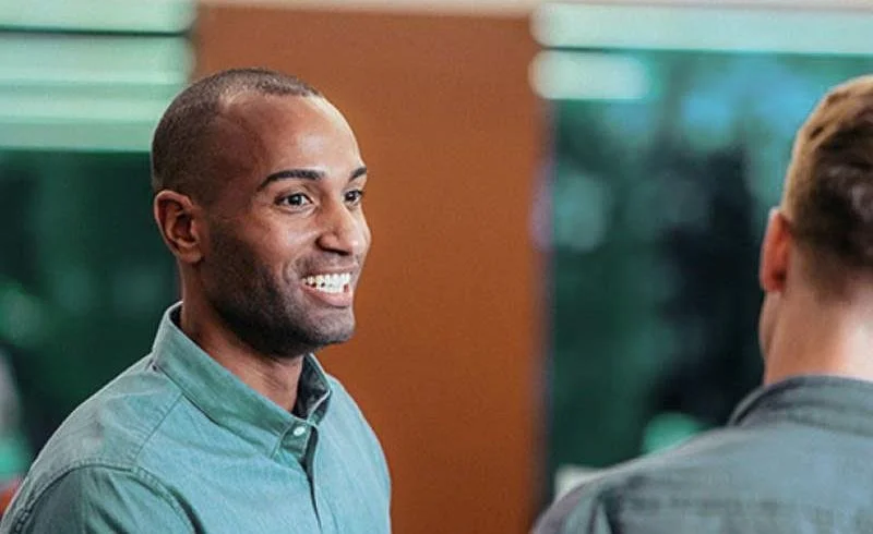 Two men talking indoors, one smiling and facing the camera, the other with his back to the camera, both in casual shirts.