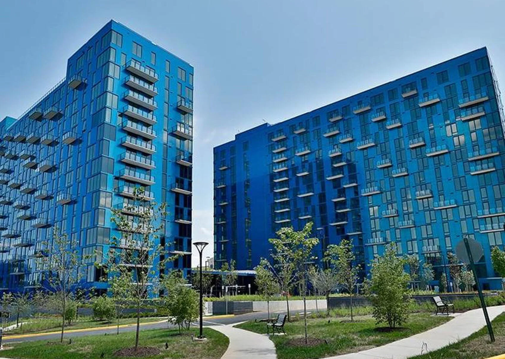 Two modern blue high-rise apartment buildings with multiple balconies, surrounded by a landscaped park with pathways, benches, and young trees.
