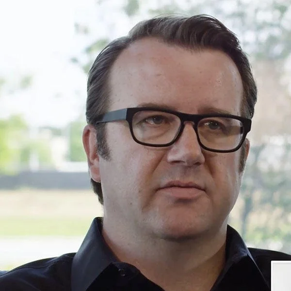 Close-up of a man with dark hair, wearing glasses, in front of a blurred outdoor background.